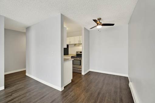 A room with a ceiling fan and a kitchen area in the background.