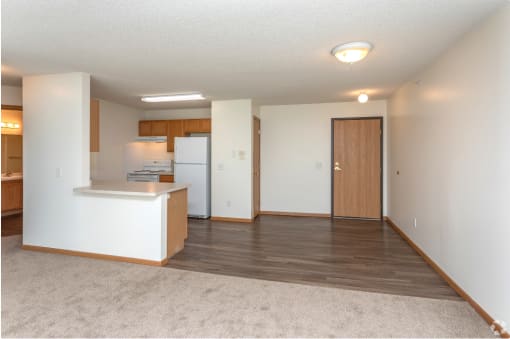 an empty living room with a kitchen and a door to a refrigerator