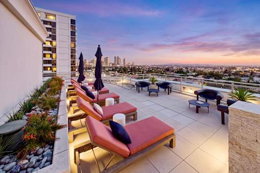 A patio with red cushioned chairs and umbrellas overlooking a cityscape at dusk.