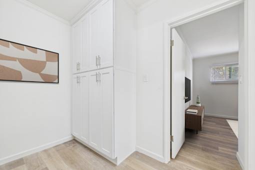 A room with a brown and white abstract painting on the wall, a white cabinet with a door, and a small table with a vase and a book on it.