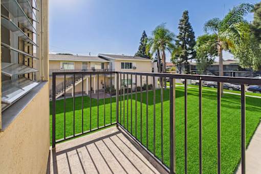 A balcony with a black railing overlooks a green lawn and a row of houses.