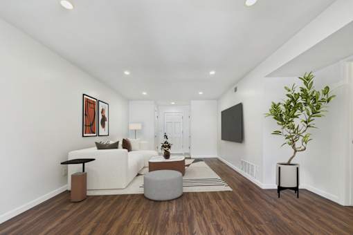 A living room with a white couch, a grey ottoman, a wooden floor, a television, and a potted plant.