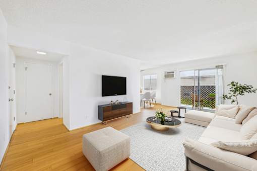 A living room with a white couch, a white chair, a white ottoman, a black television, a wooden floor, and a white wall.