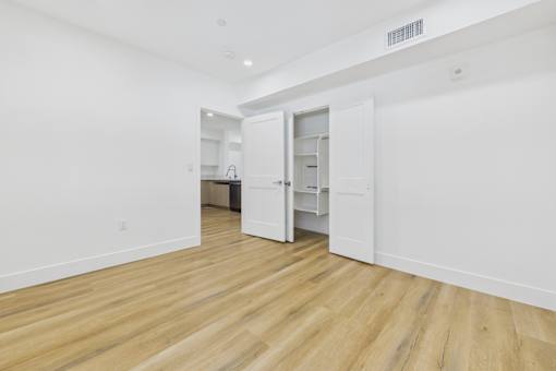A room with white walls and wooden flooring with a kitchen in the background.
