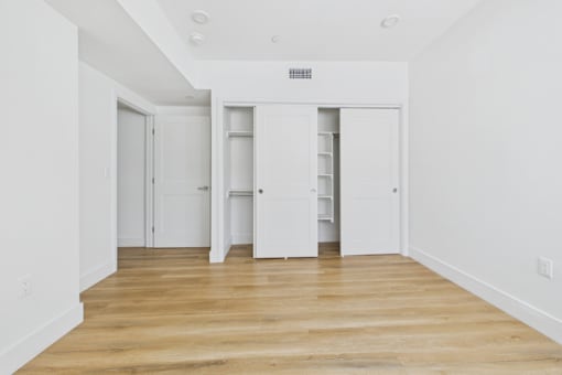 A room with white walls and wooden flooring with a white door on the left and a white cabinet with shelves on the right.