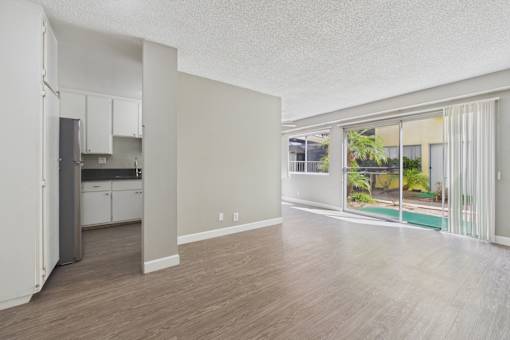 A spacious kitchen with white cabinets and a refrigerator is adjacent to a living room with a sliding glass door leading to a balcony.