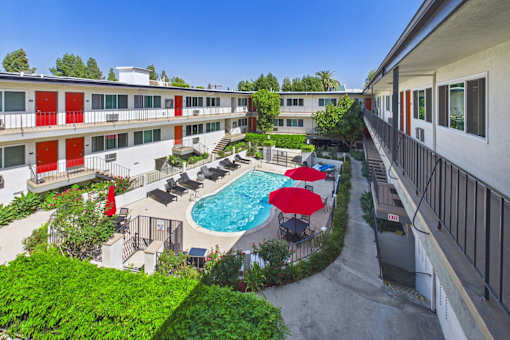 A pool surrounded by red umbrellas and chairs is surrounded by apartment buildings.