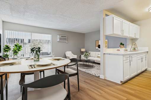 A dining room with a table set for two and a kitchen area with white cabinets.