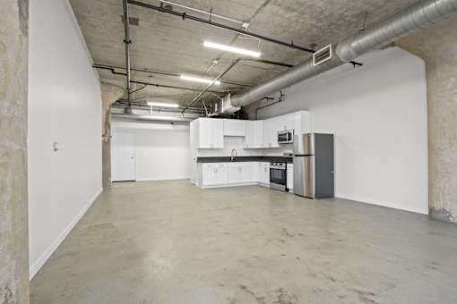 A large empty room with a kitchenette and a stainless steel refrigerator.