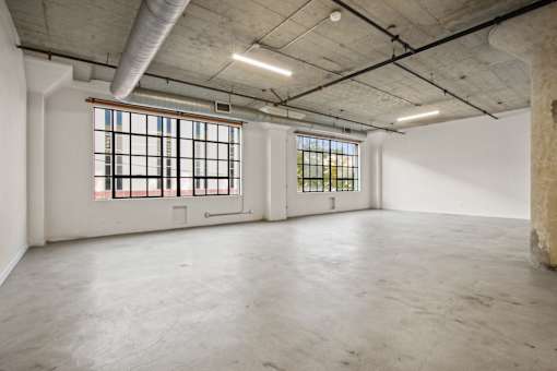 A large empty room with concrete floors and walls, two windows with bars, and exposed ceiling beams.