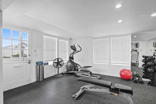 A room with a row of windows covered in blinds, a fan, a rowing machine, and a red ball.