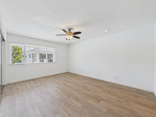 A room with a ceiling fan and light fixture, a window with blinds, and wooden flooring.