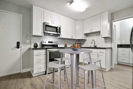 Open kitchen with stainless steel electric stove with micro hood microwave, tile backsplash, white kitchen cabinets, gray quartz counter tops, bathroom on the right