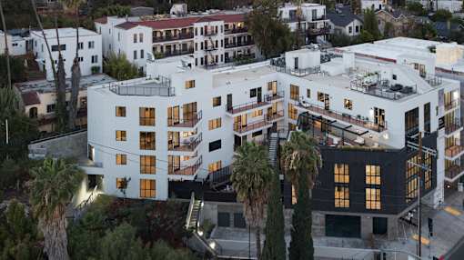 A white building with black windows and balconies is surrounded by palm trees.