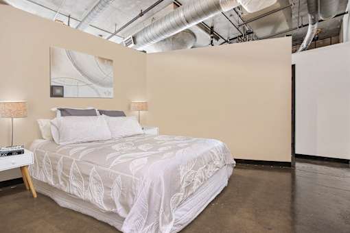 A bed with a white comforter and two pillows sits in a room with a concrete floor and a white wall with a painting and a lamp.
