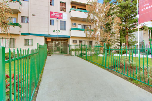 an apartment building with a green fence and a grey walkway leading to the front door