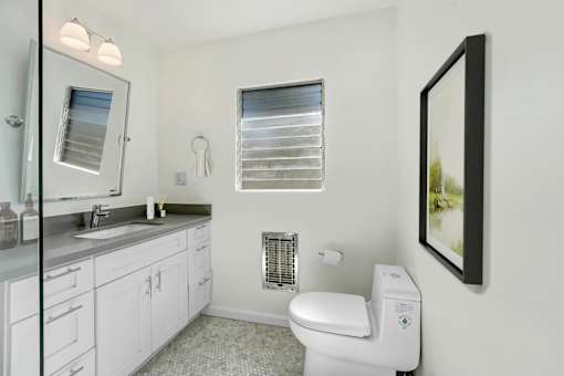 A white toilet is in a bathroom with a white sink and a window with blinds.