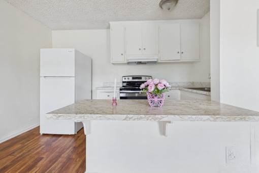 A kitchen with a white fridge and cabinets, a marble countertop, and a wooden floor.