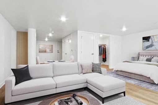 A white couch with a black pillow is in the middle of a room with a bed in the background.