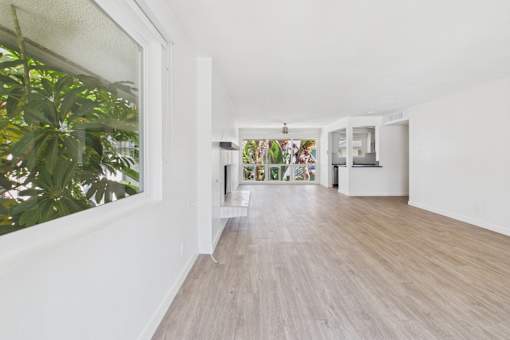 A long hallway with a white wall and a window with a view of a green plant.