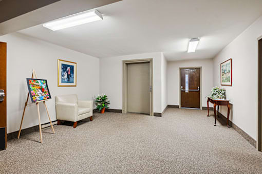 A room with a carpeted floor, a chair, a small table, and a painting on the wall.