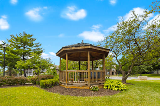 A wooden gazebo is surrounded by green grass and trees.