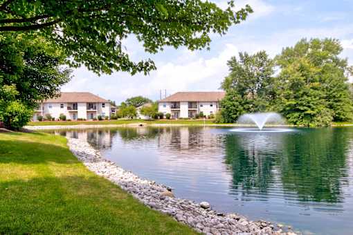 A serene lake with a fountain in the middle of it surrounded by green trees and buildings in the background.