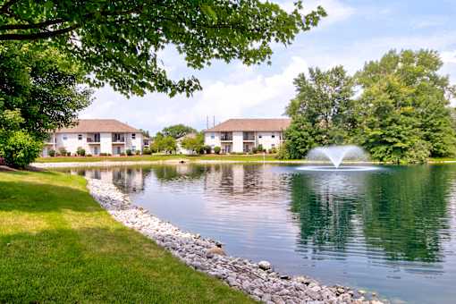 A serene lake with a fountain in the middle of it surrounded by green trees and buildings in the background.