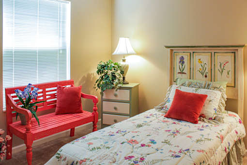 A red bench with a vase of flowers sits next to a bed with a floral comforter.
