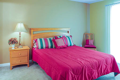 A bed with a pink comforter and striped pillows sits in a room with a wooden nightstand and lamp.