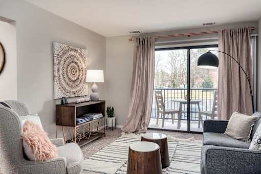 A living room with a grey sofa, a grey armchair, a wooden side table, a wooden coffee table, a lamp, a rug, and a large window with curtains.
