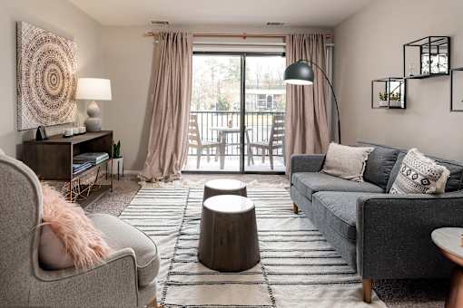 A living room with a grey couch, a grey armchair, a wooden side table with a lamp, a round wooden table, a rug, and a large window with curtains.
