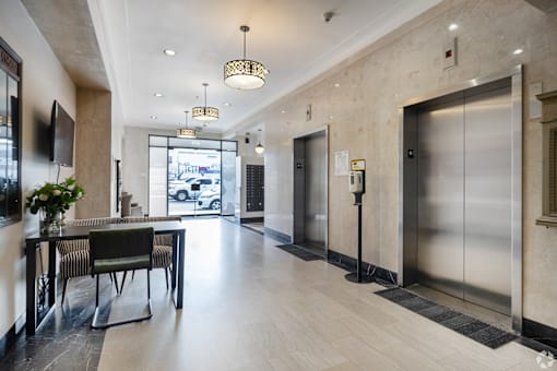 A long hallway with a table and chairs on the left and two elevators on the right.