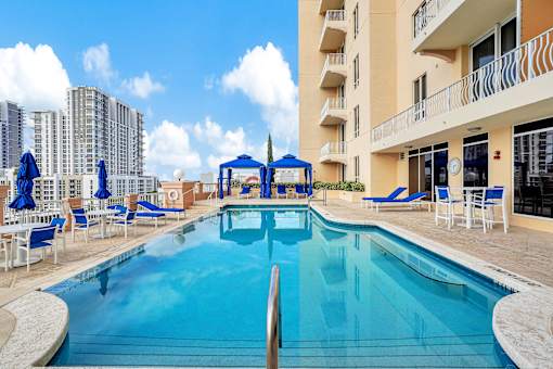 A large swimming pool in front of a building with blue umbrellas and chairs.