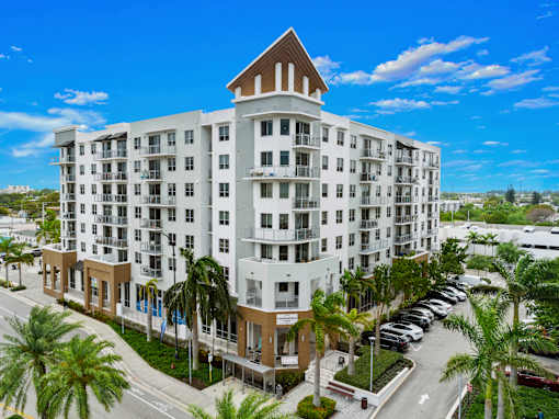A large white building with a brown roof and balconies is surrounded by palm trees.