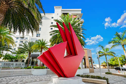 A large red sculpture in the foreground with a white building and palm trees in the background.