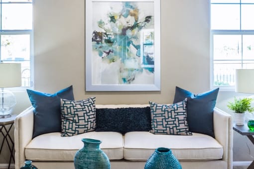 a living room with a couch and pillows and a painting on the wall flanked by windows