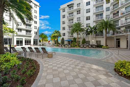 A swimming pool surrounded by palm trees and lounge chairs in a sunny courtyard.