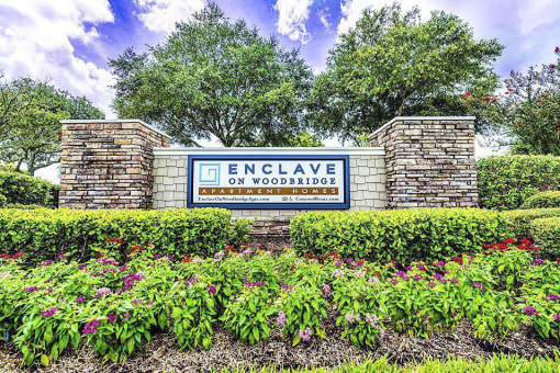 Stone pillar sign with a blue framed insert reading Enclave on Woodbridge Apartment Homes. The sign is surrounded by neatly manicured shrubs and flowers, in front of mature trees.