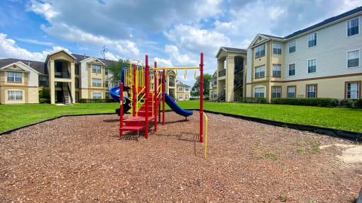 Yellow and Red Playground set with two blue slides in a bed of mulch with buildings and tree in the background