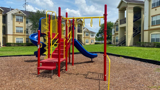 Yellow and Red Playground set with two blue slides in a bed of mulch with buildings and tree in the background