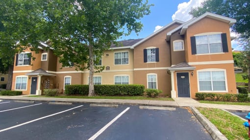Building exterior with a parking lot view surrounded by bushes and trees and  a side walk.