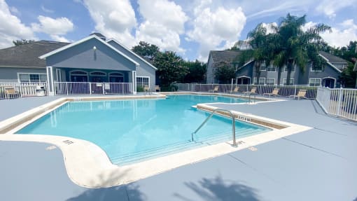 Community pool with sundeck, lounge chairs, with lake view surrounded by white metal fence with palm trees and building exteriors, palm trees, and trees in the background
