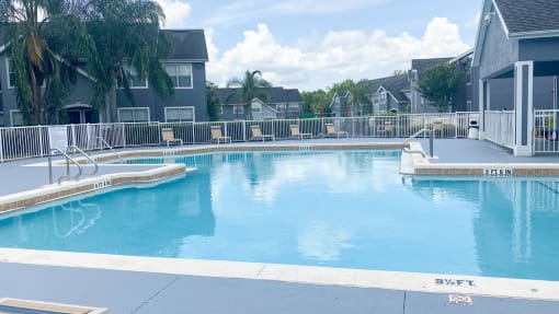 Community pool with sundeck, lounge chairs, with lake view surrounded by white metal fence with palm trees and building exteriors, palm trees, and trees in the background