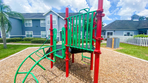 Green and Red Playground set in a bed of mulch with buildings and palm tree in the background