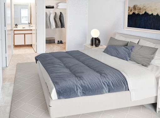 A bed with a blue comforter is in the foreground of a room with a wardrobe and a painting on the wall.
