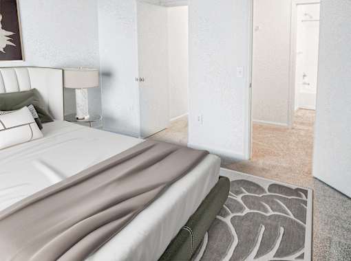 A bed with a white headboard and a grey blanket is in the foreground of a room with a lamp and a pillow on the headboard.