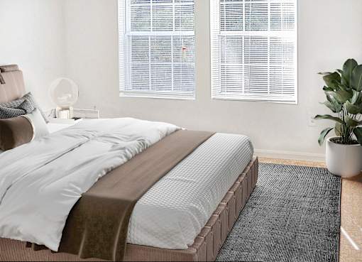 A bed with a white comforter and a brown blanket is in the foreground of a room with a fan and a plant.