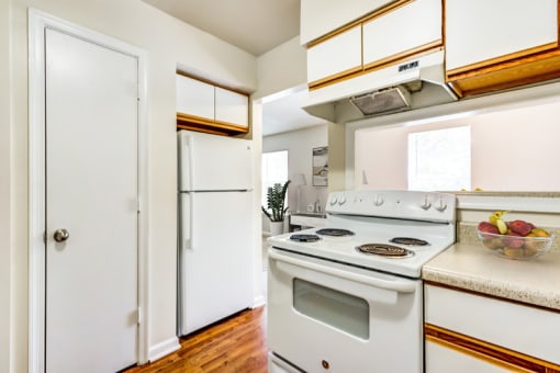 A vacant kitchen with white walls, hardwood style flooring, light countertops, a pantry door, white cabinets with light wood trim, and white appliances including a refrigerator and a stove/oven combo. There is a breakfast bar located behind the stove, and a bowl of fruit sits on the countertop.