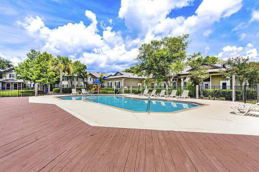 Irregular-shaped swimming pool and sundeck with tables, chairs, and loungers. The perimeter fence is lined with trees and the exterior of the clubhouse.